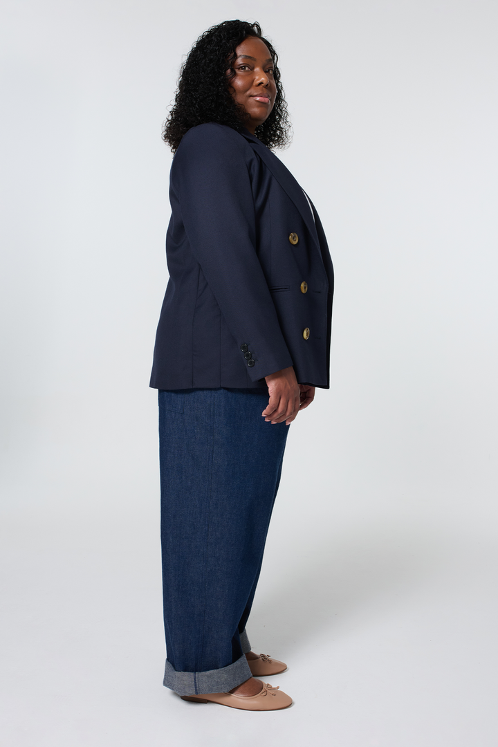 A woman in a blue adjustable blazer & wide-leg jeans standing sideways against a plain background.