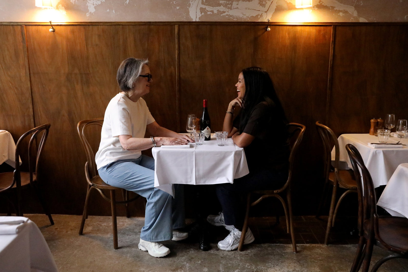 Two women sitting at a small table in a restaurant, having a conversation. The table is covered with a white tablecloth and has a bottle of wine, glasses, and salt and pepper shakers.