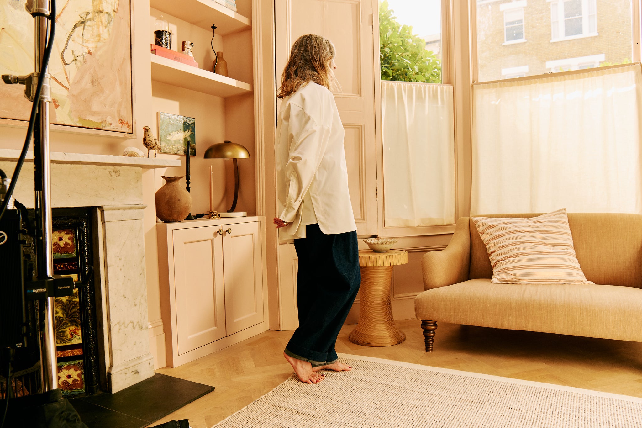 Woman in a white sustainable overshirt and adjustable wide-leg jeans standing in a stylish living room with a fireplace, modern décor, and neutral tones.