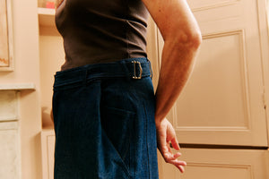 Woman wearing a brown tank top and wide-leg adjustable blue jeans made with conscious design, standing in a neutral beige-toned room. Sustainable denim that combines comfort, versatility, and timeless style.