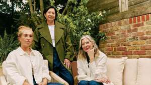 Three women wearing adjustable clothing, sustainable clothing, sustainable trenchcoat, sustainable overshirt, sustainable wide-cut jeans, sitting outdoors with greenery and a brick wall in the background.