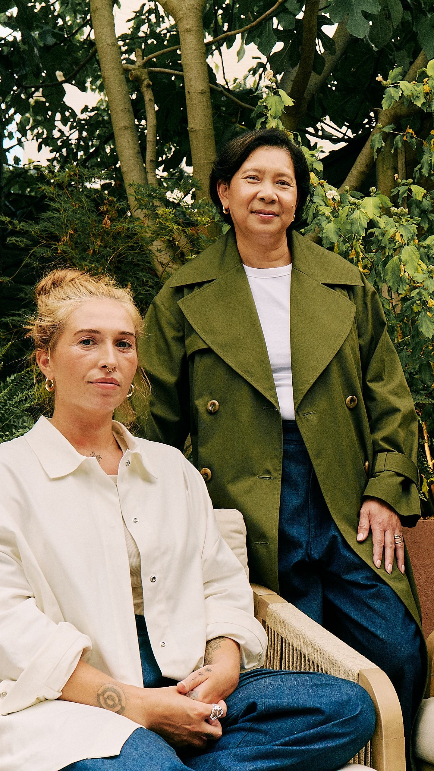 Three women wearing adjustable clothing, sustainable clothing, sustainable trenchcoat, sustainable overshirt, sustainable wide-cut jeans, sitting outdoors with greenery and a brick wall in the background.